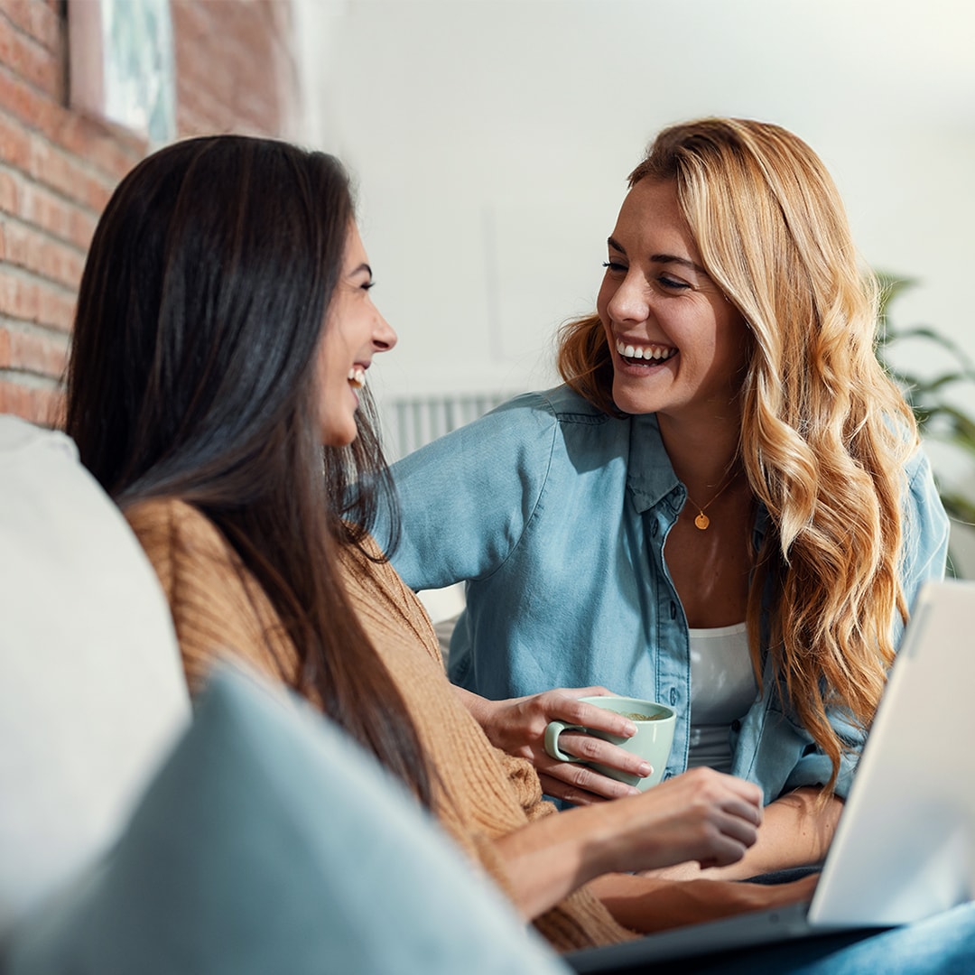 Twee vrouwen zitten samen lachend op een bank. De een houdt een mok vast, terwijl de ander een laptop op schoot heeft. 